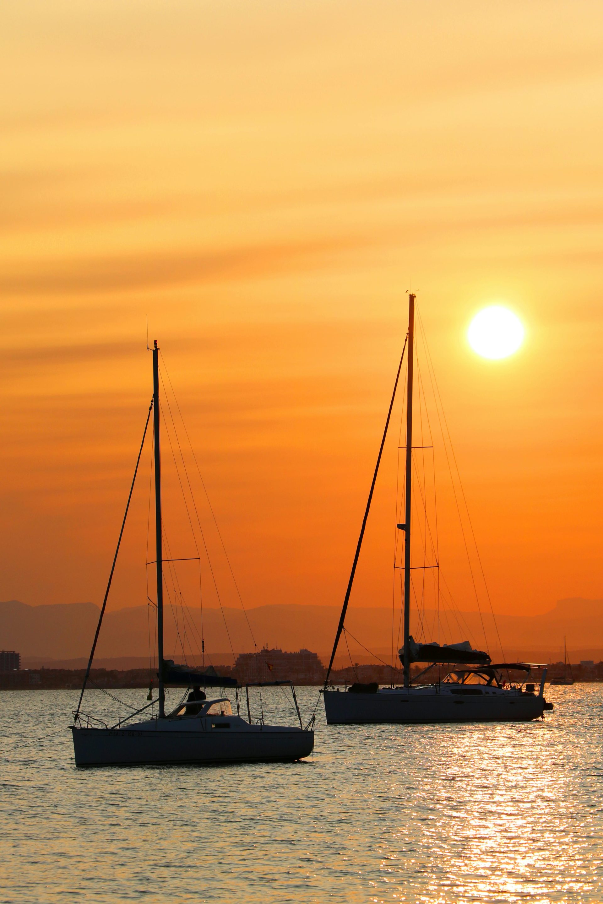 Serene scene of sailboats on the Costa Brava at sunset with a vivid orange sky.